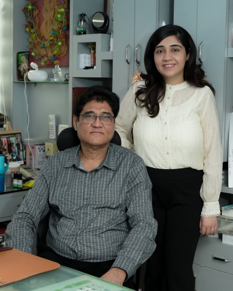 Dr. Dilip Hemnani and Dr. Sonal Hemnani standing together in their clinic, with Dr. Dilip sitting at his desk.