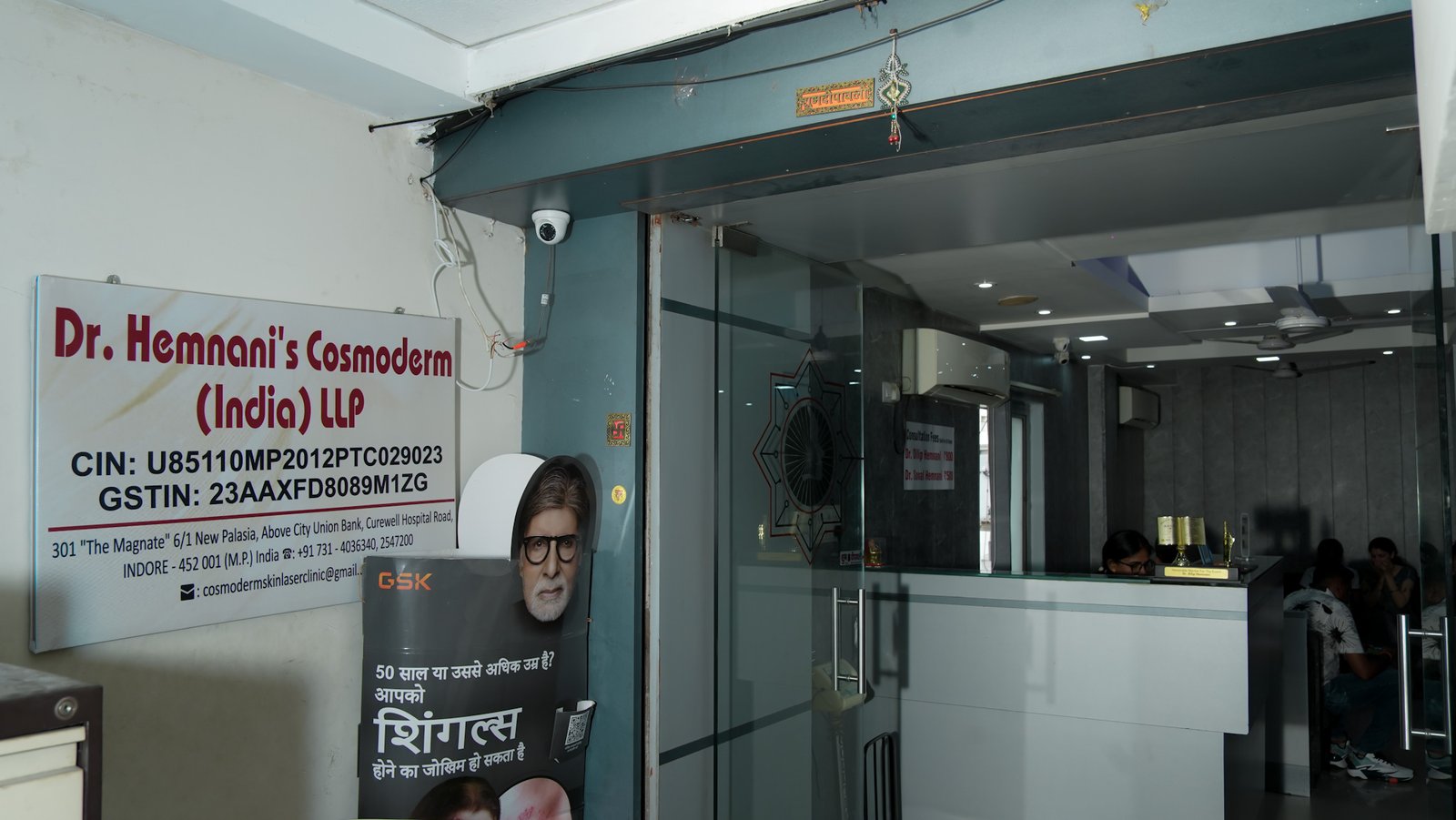 Interior view of Dr. Hemnani’s Cosmoderm (India) LLP clinic showing the reception area with a white counter, glass doors, and patients seated inside. A signboard displaying the clinic’s name and contact information is visible on the left wall.