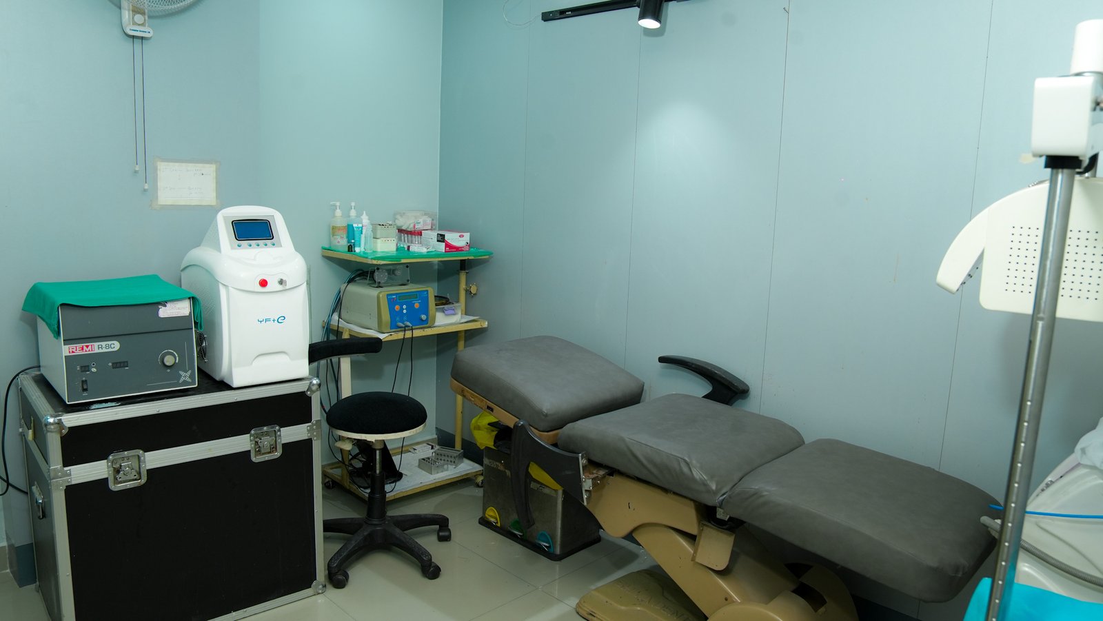 A clean medical examination room featuring a gray reclining examination chair with adjustable sections. Next to it is a black stool and a metal case topped with medical devices, including a white machine with a digital screen. Shelves in the corner hold bottles of sanitizer and other medical supplies. The walls are light blue, and the floor is tiled.