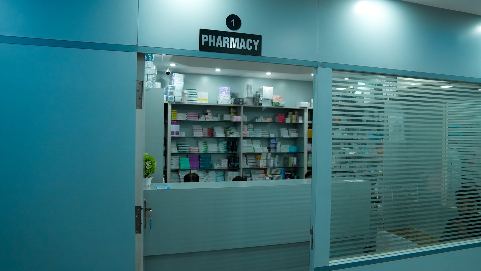The pharmacy section of a clinic is seen through a glass window with frosted horizontal stripes. Inside, shelves fully stocked with boxes of medicines line the back wall. The pharmacy counter is in front with three people partially visible behind it. The door is open and a black sign with white text above it reads 
