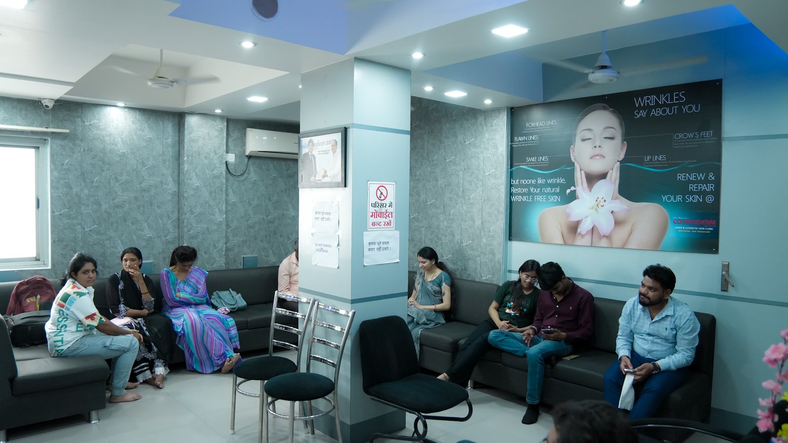The waiting room of Dr. Hemnani’s Cosmoderm clinic shows patients seated on sofas arranged around the room. The walls have grey textured wallpaper, and a large poster highlights wrinkle treatment services. The atmosphere is calm and well-lit with recessed lighting.