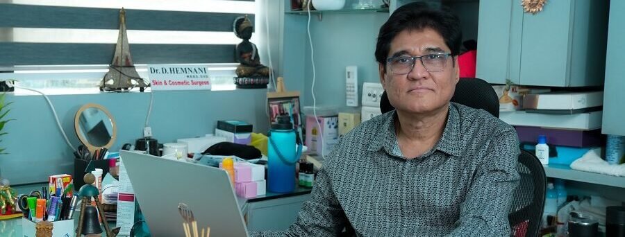 A middle-aged male doctor wearing glasses and a patterned shirt sits at his desk in a well-organized medical office with a laptop and various medical supplies around him.