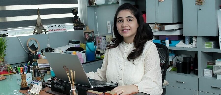 Dr. Sonal Hemnani sitting at her clinic desk, smiling, with a laptop in front of her and shelves with skincare products in the background.